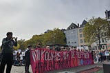 A magenta FFF Cologne banner “From Cologne to the Philippines” held up by local schoolchildren under grey skies on the Cologne Heumarkt for the Global Climate Strike 23.9.2022. The banner names the cooperation with YACAP — “Youth Advocates for Climate Action Philippines” and the hashtag #PeopleNotProfit, in the foreground and background grown-up attendants of the climate strike show their support with applause and signs.