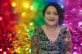 A glittery rainbow color background contains an image of Bela on the right. She is a light-skinned Latina with short, dark, curly hair and blue green eyes. She is wearing a black and white dress and has tattooed sleeves.