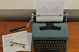 Photo of a slightly battered Olivetti Lettera 32 typewriter on a wooden table with an open A6 notebook next to it with glasses, mechanical pencil, and fountain pen