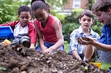 Four children playing with soil, a girl digs her hands in while a boy looks at the soil in his hands with delight