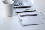 Photo of a laptop, notepad and pencil with paperclip, ruler, and mug on a white table.