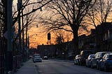 A street scene at dusk with a clear sky transitioning from orange to blue. The street is lined with parked cars on both sides and a few cars driving down the road. Bare trees with no leaves are visible, and streetlights hang from wires above the street. There are houses and buildings along the sides of the street, and a stop sign is visible on the right side of the image. The image captures the calm and quiet atmosphere of a residential neighborhood during sunset.