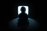 A kid sitting in front of a tv screen with flashing light in a dark room. He is so focused on it as he lose track of time he spents on it.