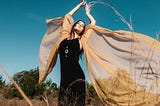 A young person with long dark hair stands with their arms held high in a field with a translucent shawl billowing around in the light breeze like wings
