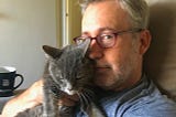A man wearing red glasses snuggles with a gray cat.