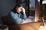Person sitting at a desk with a headset, eyes closed, holding a tissue to their forehead, looking exhausted while working at a computer.