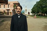 Colonel Aureliano Buendía stands in the town square of Macondo, wearing formal 19th-century black attire, with colonial buildings and villagers in the background. Scene from Netflix’s One Hundred Years of Solitude series.