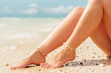 Bare legs of a woman sitting on a beach wearing nothing but ankle bracelets.