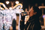 Pleasant brown haired tanned caucasian woman drinking takeout coffee on street corner at night with festive clear white winter lights