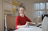 A previously stern woman sits at her desk smiling broadly.