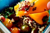 Tupperware box with quinoa salad, tomatoes, and a hand holding a small bell pepper with some salad on it as a spoon.