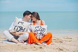 A young couple sitting on a beach with the sea in the background. He is wearing white and holding a cushion that reads Mr right. She is wearing a bright orange dress and holding a cushion that reads Mrs always right.