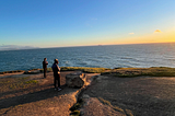 My friends overlooking the beautiful California coast. They’re on a rock cliff staring at the sunset starting to overtake the ocean.