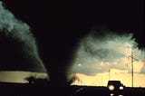 [Image: black tornado close behind a moving vehicle, the sky cloudy and foreboding in the background.]