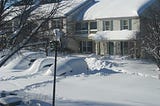 In the front left, you can see snow-covered branches of a tree jutting out, with a fancy light on a pole to the right. The photo shows cars on both sides of the unplowed street. They are so covered by snow that it’s hard to tell them apart. In the background, you can see snow-covered lawns and houses with roofs topped by heavy snow.