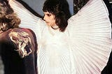 Freddie Mercury wearing a ruffled, white cloak in a slightly dim room.