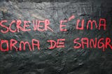 Um muro pintado de preto contém uma pichação em vermelho, escrito “escrever é uma forma de sangrar”