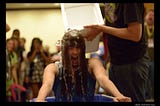 A woman is seen yelling while a bucket of ice water is being poured over her head. She’s in the middle of the room surrounded by dozens of people in the background, out of focus, seemingly cheering her on.