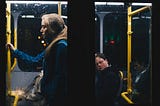 A blonde lady, in her twenties, stands on a bus. She is being watched by men around her.