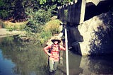 Woman wearing a hat standing in a creek in front of a concrete structure .