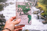 A man’s hand holding a card that says ‘Adventure Awaits’ against the backdrop of a waterfall.