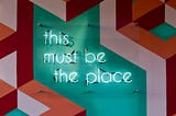 a neon sign on a colourful wall, saying “this must be the place”