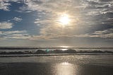 Image of a beach with ocean waves crashing. The sky is blue and the sun is shining through the clouds.