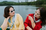 Hey middle age? You look fabulous. Two women in silly sunglasses and cute sweaters glam it up by a lake.