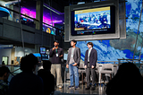 Three students stand on a stage with a large monitor behind them displaying a group of people and the words “Thank you.”