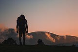 A silhouette of a person trekking towards an orange sky and large mountains.
