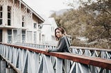 A slender, fair-skinned lady stands on a bridge with her folded hands placed on the metal and wood rails of the bridge asooks on in deep thoughts.