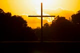 A brown cross above the water in front of a yellow sky.