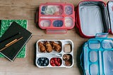 Photo of pink and blue lunch boxes and trays on table next to a green and black notebook and pencils.