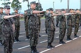 Soldados do Exército brasileiro fardados fazem o gesto de juramento à bandeira nacional, com o braço erguido horizontalmente, em posição paralela ao chão