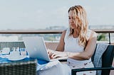 Image of a woman working on her laptop at a table on a balcony overlooking a cityscape