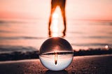 A figure standing on a glass ball as the sun sets on the beach
