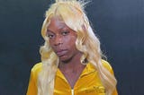head and shoulders photo of a black woman wearing a gold blouse. she has long blond hair past her shoulders. She is not smiling, but she is looking at the camera
