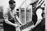 Two men from the 1940s discuss the farm products one of them is holding