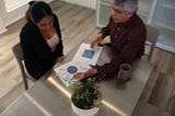 An overhead image of two people sitting at a conference table, with one person showing two charts that are printed on two pieces of paper and explaining the significance of the charts to the other person.