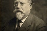 A portrait of Rudolf Rocker. In this photo, he has oval-shaped, wire glasses, a white mustache and goatee, and salt-and-pepper, slightly curly hair. He’s wearing a suit with a vest and a tie.