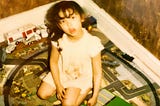 Little girl with dark hair sitting in the middle of a train set looking up at the camera.