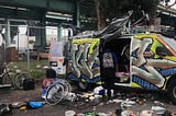 A figure stands in the open door of a gratified van, surrounded by bike parts, a cooler, and other supplies