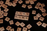 scrabble tiles spelling out “Stay Home” on a black background, with other scrabble letter tiles scattered around it