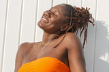 Black woman with locs smiling under sun in orange top