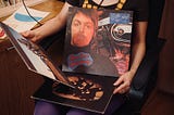 Seated female teenager in 1970’s clothes with albums in her lap one of Paul Mccartney