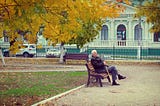 An elderly man sitting on a park bench. He is wearing an overcoat and holding a walking stick. The yellow and gold leaves of two trees are visible in shot, and fallen leaves litter the ground. There are no other people visible in the park.