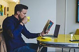 A man reading a book at a yellow table while drinking from a yellow cup.