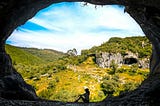 A young woman sits at the entrance of a dark cave. Beyond the entrance, a green valley is lit by bright sunshine from a blue sky which shows only wisps of white cloud. Across the valley, another, similar cave entrance can be seen in a limestone outcrop.