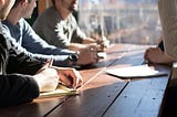 A tight shot of a wooden table in front of a large window, through which a city skyline can be seen. At least four people are sitting around the table, because their arms can be seen resting on it. Thrree people are holding pens and have notepads in front of them.