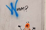 Grafitti on a white wall — “Warum?” is written in black, with the W spray-painted in blue, above an orange bean-shaped character with black, gangly limbs.  Expression is hard to read because of damage to the wall, but eyebrows are raised off its head. Some kind of bricks or brick-effect cladding at the bottom of the image give the impression of a small drawing (maybe 20cm high?) at the bottom corner of a wall.